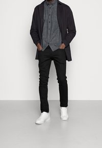 Zwarte jeans, een grijs overhemd met korte mouwen en een ruitpatroon, en een donkere jas, aangevuld met witte sneakers.