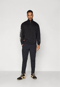 Černá sportovní mikina s vysokým límcem a zipem, s potiskem na rukávu. Kombinována s tmavými kalhotami a černou obuví s béžovými akcenty.