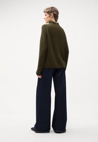 Maglione a maglia verde oliva con collo alto, abbinato a jeans di denim scuri a gamba larga, visto da dietro, che mette in evidenza la vestibilità rilassata.