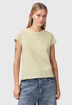 Ung kvinde med langt blondt hår iført en lys beige T-shirt med korte ærmer og blå denimjeans, stående foran en ensfarvet baggrund.
