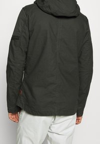 Mörkgrön huva jacka med matt yta, som har en dragkedjeficka på ärmen och en tygpatch på bakfåll.