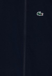 Veste zippée marine en tissu texturé avec un petit logo de crocodile vert sur le côté gauche de la poitrine.
