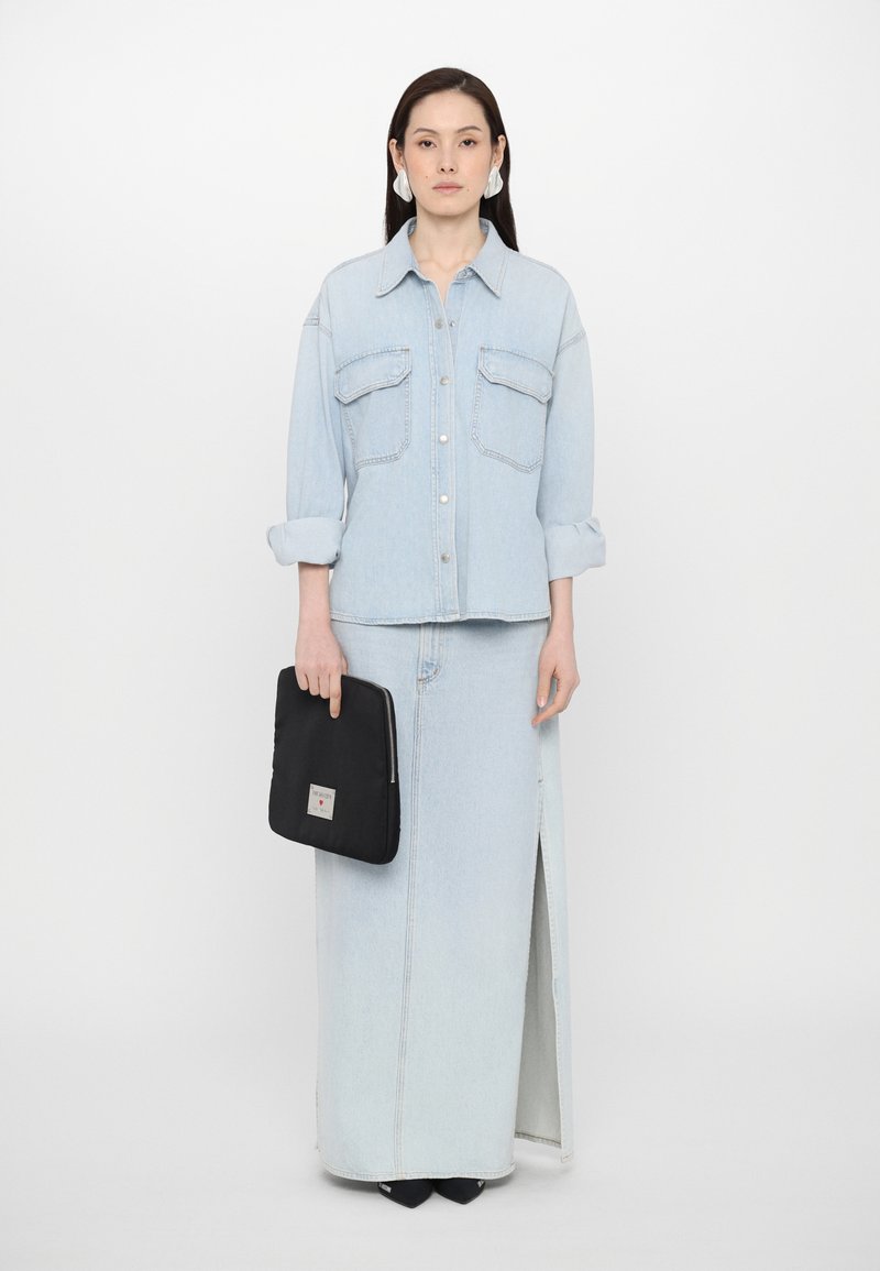Camisa de mezclilla azul claro con dos bolsillos en el pecho, sin abotonar en el cuello, combinada con una larga falda de mezclilla azul claro que presenta una abertura lateral. Clutch negro con cremallera.