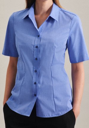 Lyseblå kortærmet skjorte med knapper og spids krave, båret af en person med hænderne i lommerne på sorte bukser.