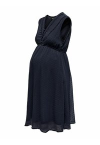 Robe de maternité sans manches bleu marine avec col en V, superposition transparente à pois et taille empire, conçue pour accueillir un ventre de femme enceinte.