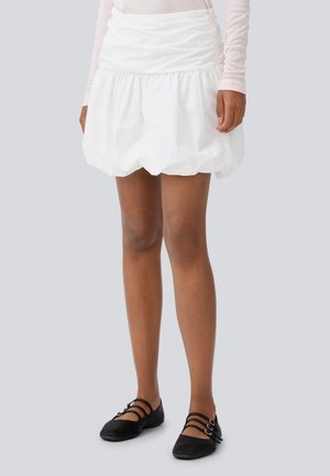 White puff skirt with a gathered waistband, featuring a textured fabric. Black flats with double straps complement the outfit.