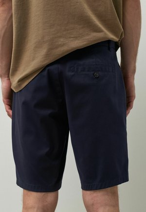 Mężczyzna ubrany w granatowe spodenki do kolan i brązowy t-shirt, pokazany od tyłu na neutralnym tle.