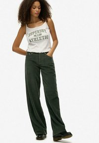 Camiseta sin mangas blanca que dice "SUPERDRY 87 ATHLETIC" en texto verde, combinada con pantalones de pana de pierna ancha de color verde oscuro y botas negras.
