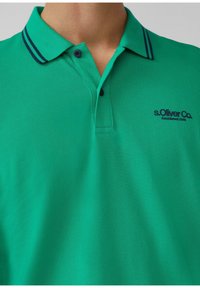 Grünes Poloshirt aus strukturiertem Stoff. Verfügt über einen Kragen, eine Zwei-Knopf-Leiste und dunkelblaue gestreifte Akzente am Kragen.