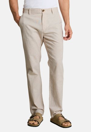 Man wearing beige straight-fit pants with button closure and beige sandals with two buckle straps, standing with one hand in pocket.