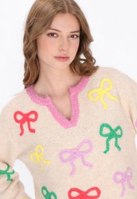 Beige Pullover mit einem rosa V-Ausschnitt, auf der Vorderseite mit bunten gestrickten Schleifen in Rot, Pink, Gelb, Grün und Lila verziert. Weicher Stoff.