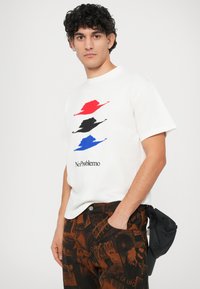 Weißes T-Shirt mit rotem, schwarzem und blauem Hut-Graphic; "No Problemo"-Text darunter. Kombiniert mit braunen gemusterten Hosen.
