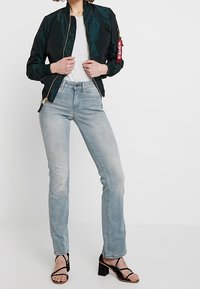 Persona con una chaqueta bomber negra, camiseta blanca, jeans de mezclilla claros y sandalias de tacón negras con tiras, de pie contra un fondo blanco.