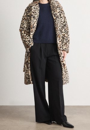 Personne portant un manteau en fausse fourrure léopard longueur genou, un pull marine, un pantalon large noir et des chaussures pointues noires, debout à l'intérieur.