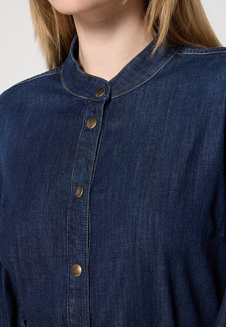 Ciemnoniebieska koszula z bandem, z pięcioma mosiężnymi guzikami z przodu i widocznymi detalami szwów. Gładka tekstura z luźnym krojem.