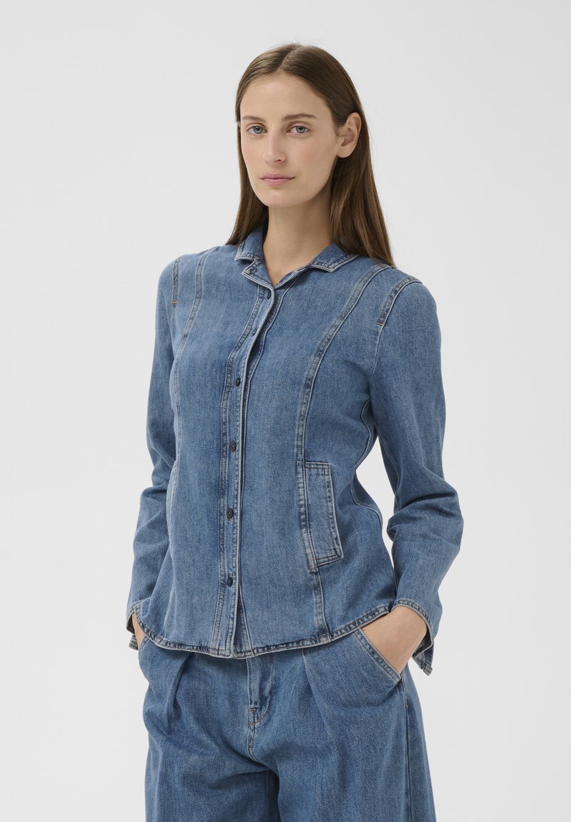 Femme aux cheveux bruns raides portant une veste boutonnée en denim bleu ajustée et un pantalon en denim bleu assorti, les mains dans les poches.