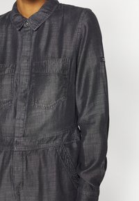 Mono gris oscuro con cuello camisero, dos bolsillos delanteros y cintura elástica. La tela parece texturizada y ligera.