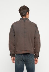 Brown denim jacket with a collar, dropped shoulders, and elastic cuffs. Features a distressed texture and a central seam at the back.