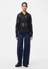Woman wearing a black sheer lace button-up blouse, dark blue wide-leg jeans, and black shoes, standing against a plain background.