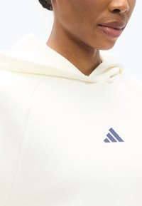 Φούτερ με κουκούλα σε κρεμ απόχρωση, με μεγάλη κουκούλα και μπλε-ναυτικό λογότυπο Adidas στο στήθος. Ύφασμα απαλό, με άνετη εφαρμογή και μανίκια ράγκλαν.