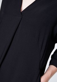 Musta paita, jossa V-pääntie, 3/4-hihat ja hienovaraiset sivuyksityiskohdat. Valmistettu sileästä, kevyestä kankaasta. Yksinkertainen, virtaviivainen muotoilu.