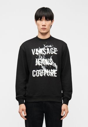 Jonge man met zwart haar, gekleed in een zwarte trui met een wit spattenontwerp en de tekst "Versace Jeans Couture" aan de voorkant.