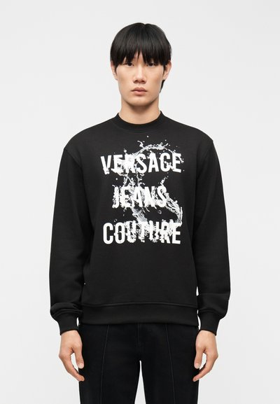 Jeune homme aux cheveux noirs portant un sweat-shirt noir avec un motif éclaboussé blanc et le texte "Versace Jeans Couture" à l'avant.