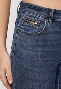 Mörkblå denimjeans med en zippered ficka, slim fit och minimala sömnadsdetaljer. Materialet verkar mjukt med en slät yta.