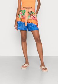 Femme portant un short à motifs tropicaux avec des palmiers et des vagues de l'océan, associé à des sandales beiges à enfiler et un haut sans manches orange.