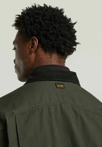 Oliven-grøn jakke med en corduroy-krave, der har et firkantet mærke på bagsiden, glat tekstur og et afslappet fit-design.
