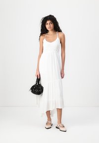 White maxi dress with thin straps, crochet detailing at the bodice, flowing hemline, and subtle texture. Paired with a black patterned handbag.