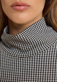 Schwarz-weißer Hahnentrittmuster-Turtleneck-Pullover mit hohem Kragen und weicher, strukturiertem Strickgewebe. Nahaufnahme.