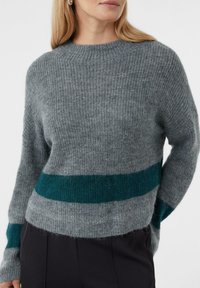 Kobieta w szarej swetrze z dzianiny z okrągłym dekoltem, z turkusowym poziomym paskiem w talii i na rękawach, w połączeniu z czarnymi spodniami.