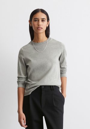 Maglietta grigia a maniche lunghe realizzata in un tessuto leggero, con scollo rotondo. Indossata con pantaloni neri, che mostrano una vestibilità rilassata.