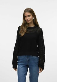 Zwarte gebreide trui met een losse pasvorm, grote mouwen en een gestructureerd open geweven ontwerp. Gecombineerd met blauwe spijkerbroek.