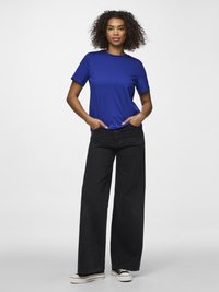 Žena stoji s rukama u džepovima, nosi plavu majicu, široke crne traperice i crno-bijele platforme tenisice na jednobojnoj bijeloj pozadini.