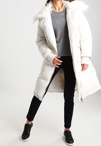 Kobieta w długim białym płaszczu puchowym z futrzanym wykończeniem, w szarym swetrze, czarnych spodniach i czarnych sneakersach, stojąca z jedną nogą wysuniętą do przodu.
