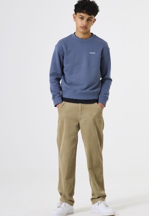 Sudadera azul con cuello redondo y pequeño logo blanco, combinada con pantalones de pana beige y zapatillas blancas, de pie sobre un fondo liso.