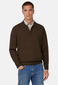 Pruun pullover-sviiter, millel on krae ja ribidega mansetid ning äär. Kandes heledat halli kraega särki, koos siniste teksadega.