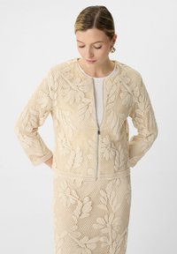 Beżowa siatkowa kurtka z kwiatowym haftem, z zapięciem na zamek i rękawami o długości trzech czwartych. Teksturowany materiał z misternymi wzorami liści.