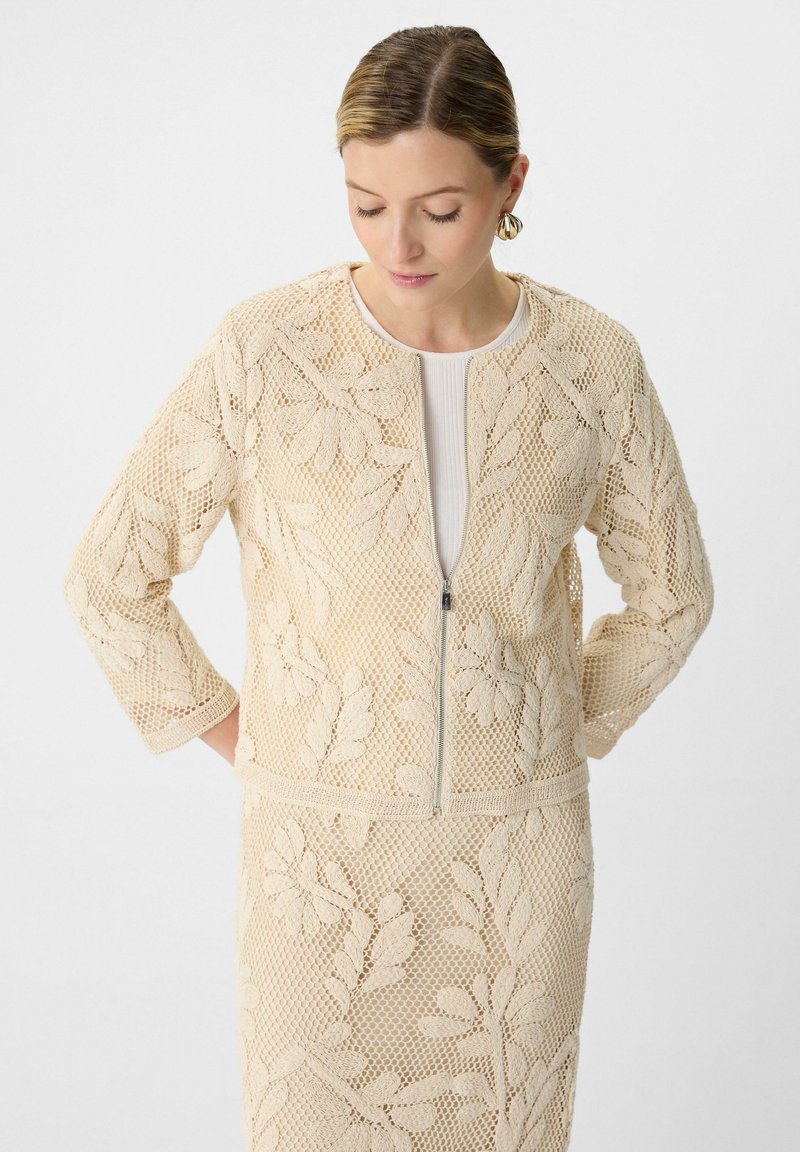 Beżowa siatkowa kurtka z kwiatowym haftem, z zapięciem na zamek i rękawami o długości trzech czwartych. Teksturowany materiał z misternymi wzorami liści.