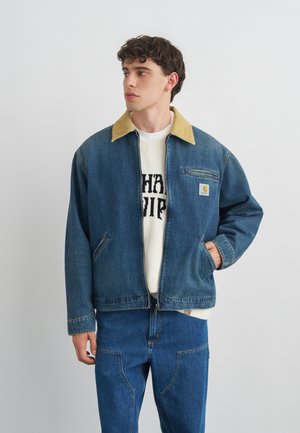 Fiatal férfi kék farmerdzsekit visel világosbarna gallérral és hozzáillő farmernadrággal, balra néz egy egyszínű háttér előtt.