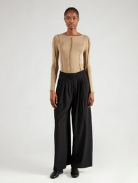 Beige Langarmtop mit einem mittigen Reißverschluss und Nahtdetails, kombiniert mit schwarzen weit geschnittenen Hosen und schwarzen Schuhen vor einem neutralen Hintergrund.