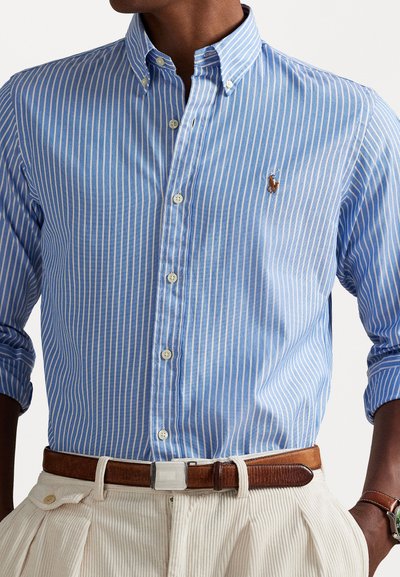Chemise à manches longues en coton à rayures bleues et blanches, avec un col. Présente un logo sur la poitrine et une fermeture à boutons à l'avant.