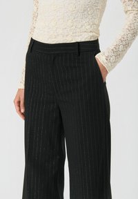 Pantalons noirs à rayures fines avec une coupe ajustée, présentant une texture lisse et des poches avant, mettant en valeur des rayures verticales raffinées.