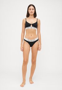 Zwart lingerie-set bestaande uit een bralette met een keyhole-uitsnijding en logo-band, gecombineerd met bijpassende slipjes. Zacht, rekbaar materiaal.