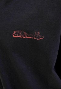 Černá bavlněná tkanina s červeným, texturovaným logem "Ellesse" v stylizované kurzívě, umístěným blízko okraje.