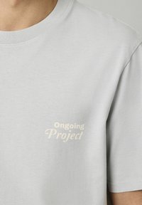 T-shirt gris clair à manches courtes fabriqué en matériau doux, arborant une impression couleur crème qui indique "Projet en cours" sur le côté gauche.