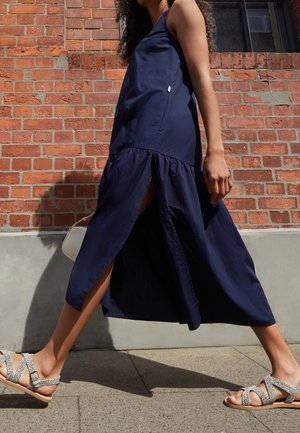 Robe maxi sans manches de couleur marine avec un ourlet à volants en tiers et des fentes latérales. Associée à des sandales claires avec un motif à taches.