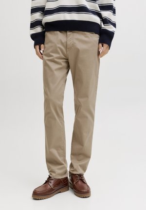 Person iført beige chinos, brune læder snørestøvler og en sort-hvid stribet langærmet sweater, stående mod en ensfarvet baggrund.
